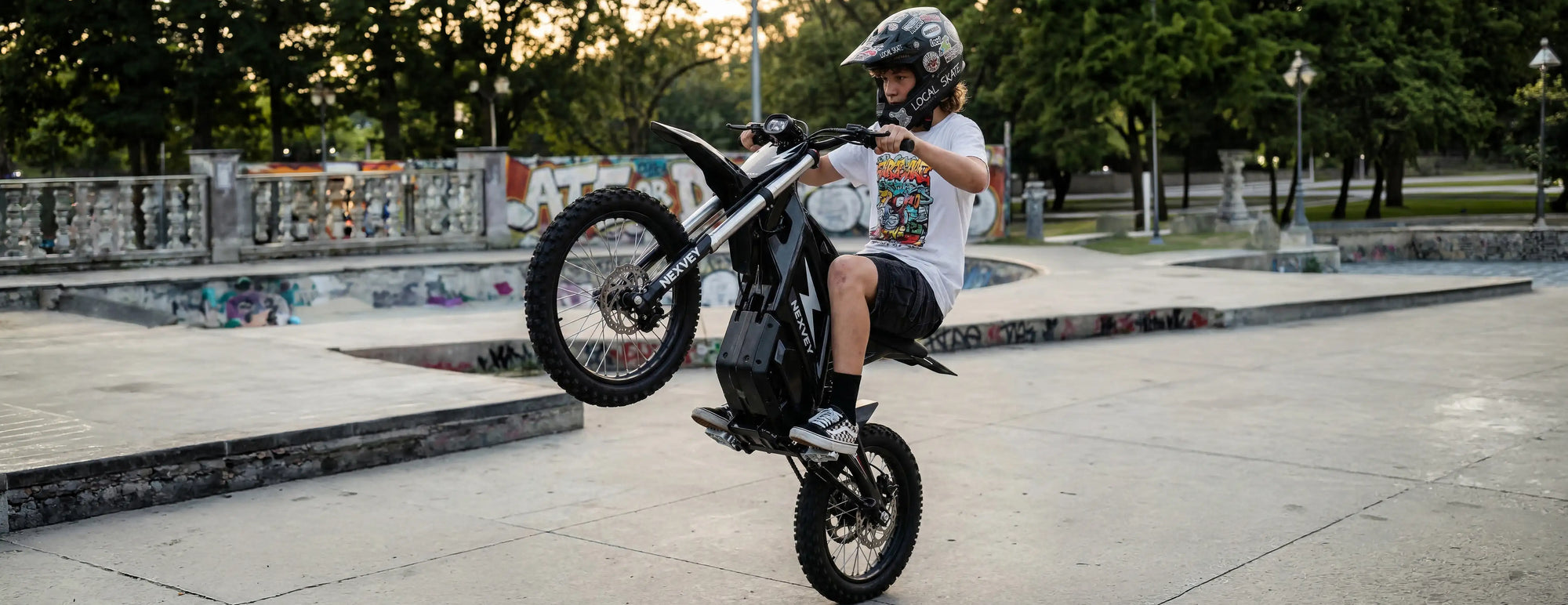 A teenager performing a wheelie stunt on a black NEXVEY electric dirt bike at a concrete skate park