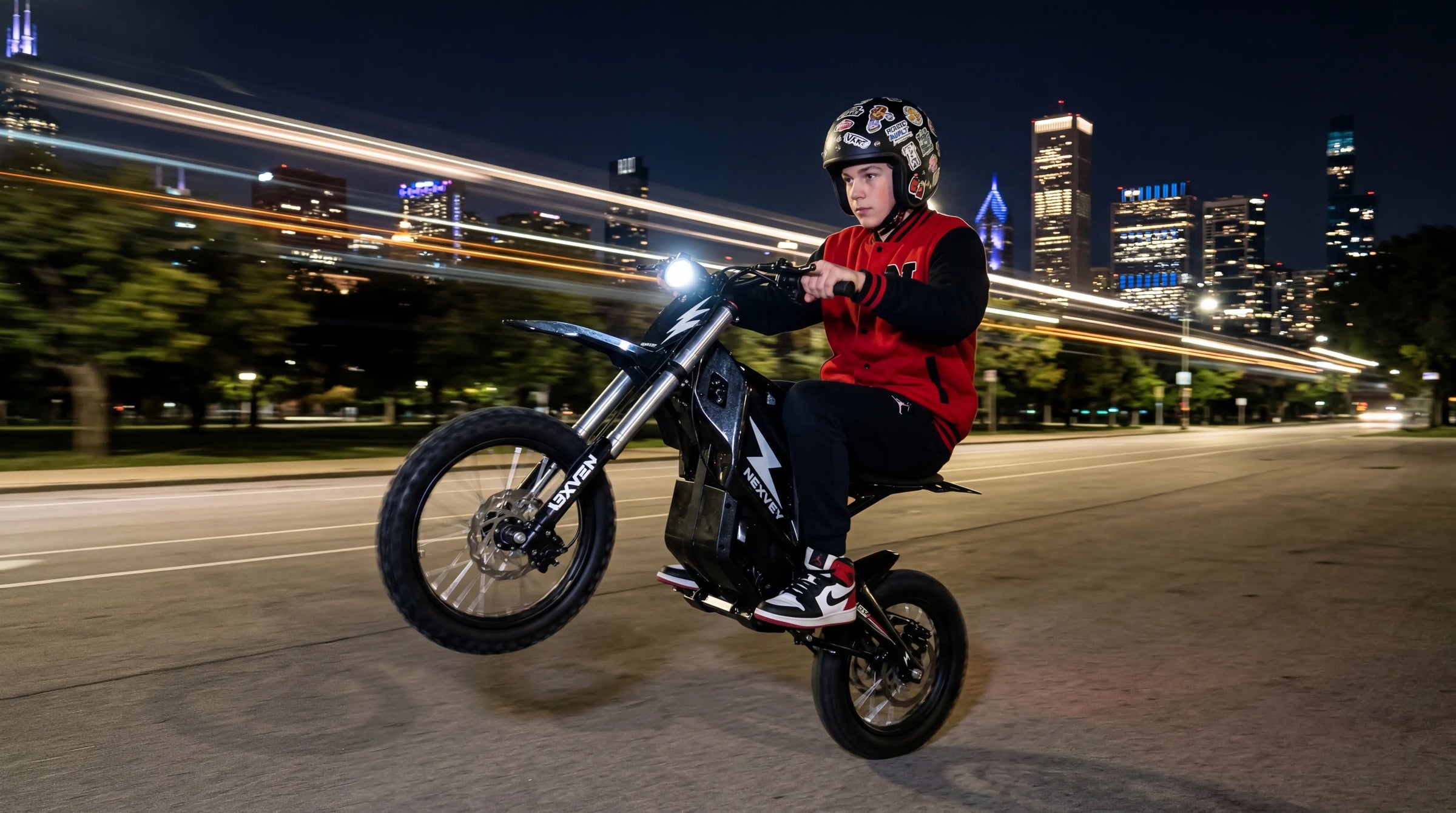 A teenager wearing a helmet and red varsity jacket performing a wheelie on a NEXVEY electric dirt bike at night with a city skyline background