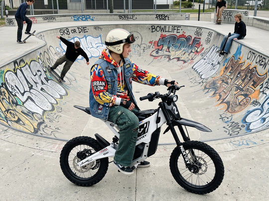 A helmeted rider on a white NEXVEY FLASH 2 electric dirt bike beside a graffiti-covered skatepark bowl