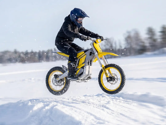 A rider in full winter gear catching air on a yellow NEXVEY FLASH electric dirt bike while jumping over a snow bank in a winter landscape
