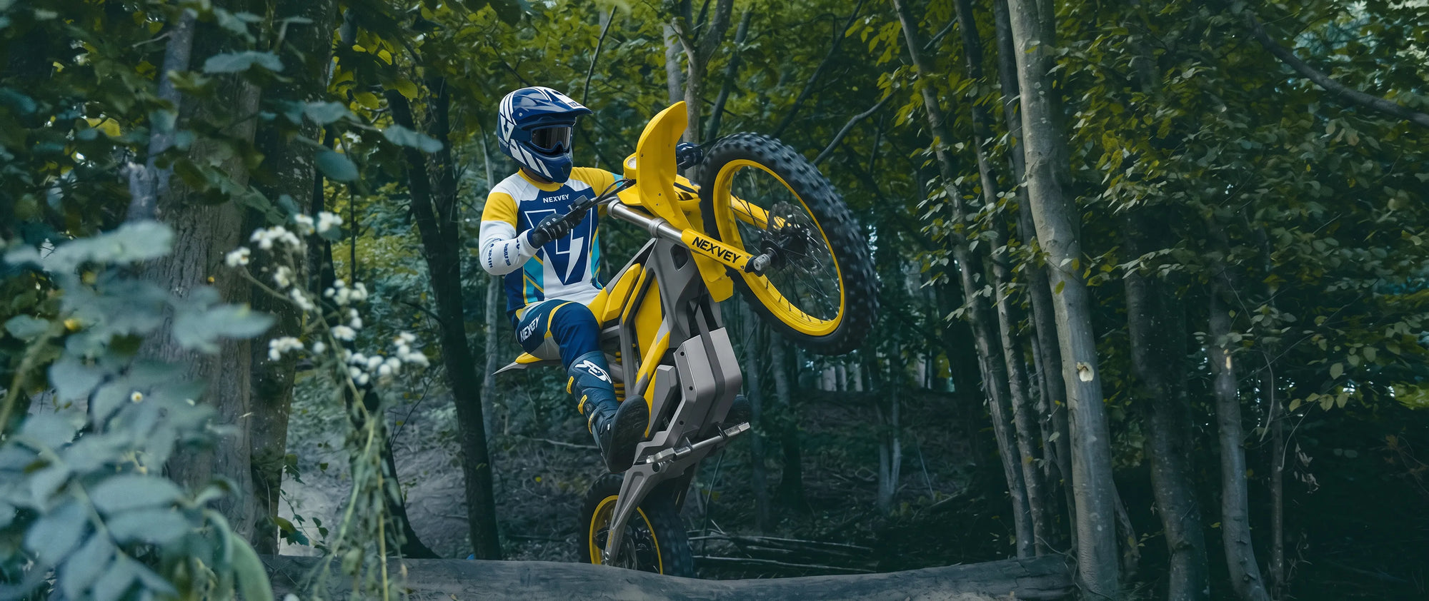 Teenager riding a yellow NEXVEY Flash electric dirt bike performing a wheelie in a wooded trail setting