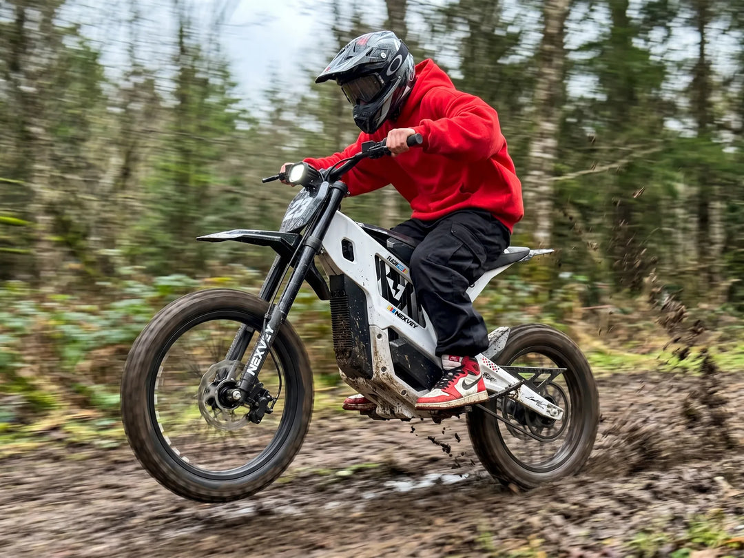 Rider in red hoodie riding the white Nexvey Flash Pro youth electric dirt bike through a muddy off-road forest trail