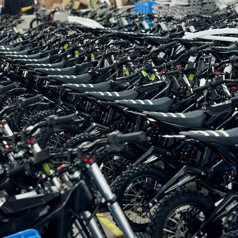 Rows of all-black NEXVEY electric flash bikes lined up in a warehouse, featuring striped seats and rugged off-road tires, highlighting mass production and ready-to-ship inventory