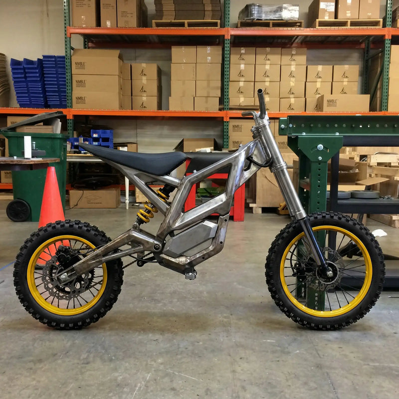 Side view of a NEXVEY electric dirt bike with a reinforced raw steel frame, yellow rear shock suspension, and knobby off-road tires with yellow rims in a warehouse setting