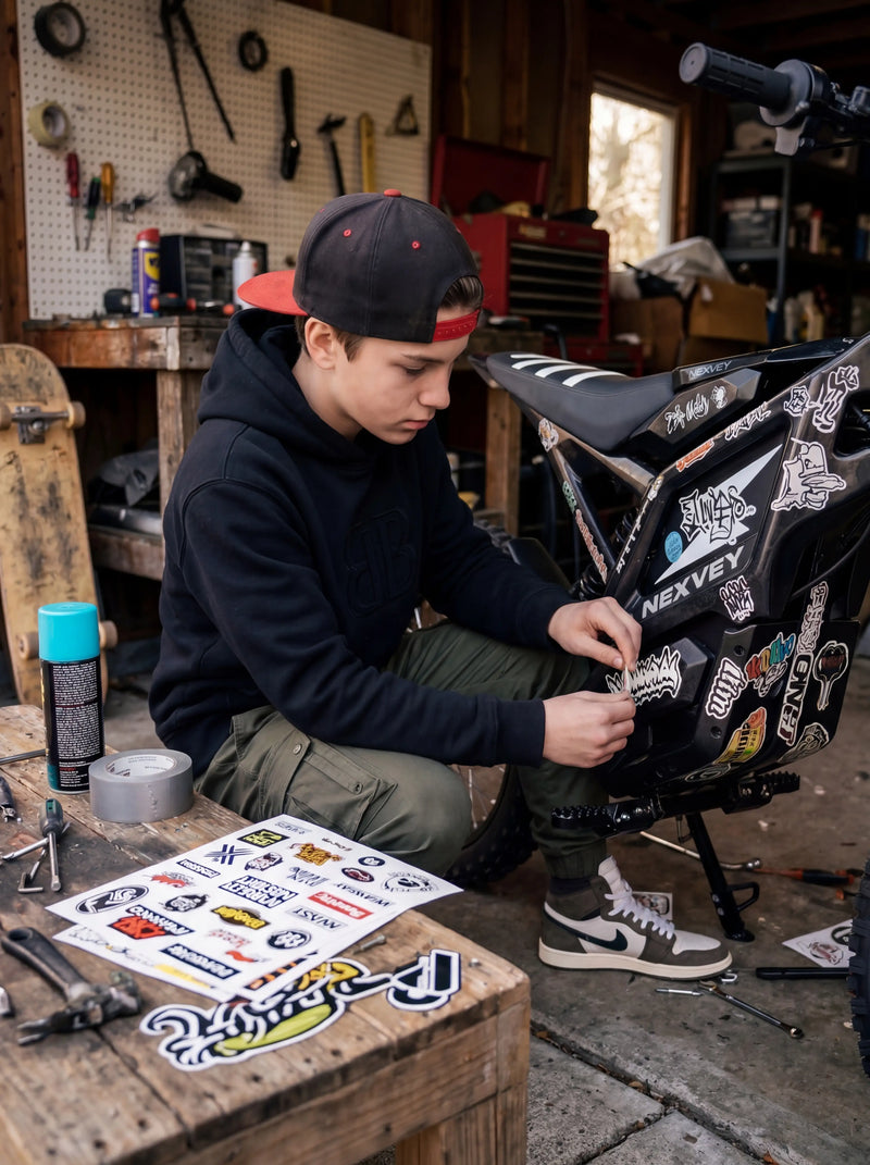 A teenage rider in a workshop carefully applying custom racing stickers to a black NEXVEY FLASH electric dirt bike, with tools and a skateboard in the background