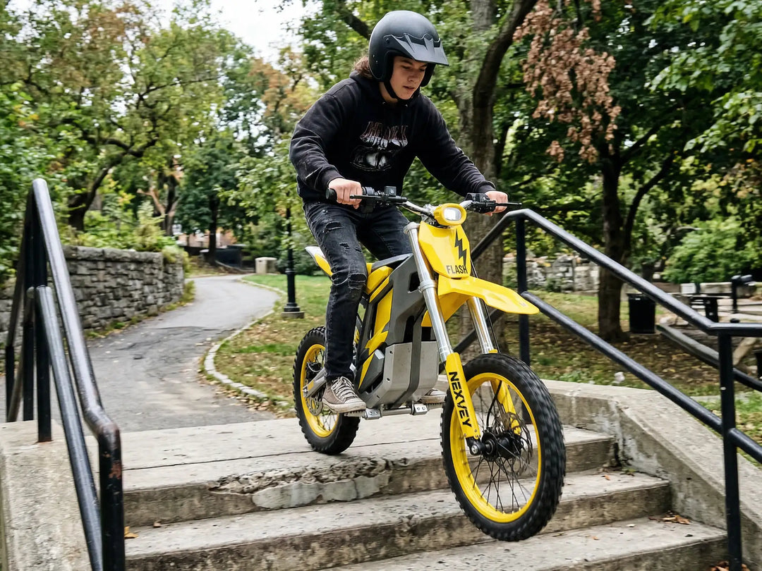 A teenage boy in a black hoodie and helmet riding a yellow NEXVEY FLASH electric dirt bike down concrete stairs in a wooded city park