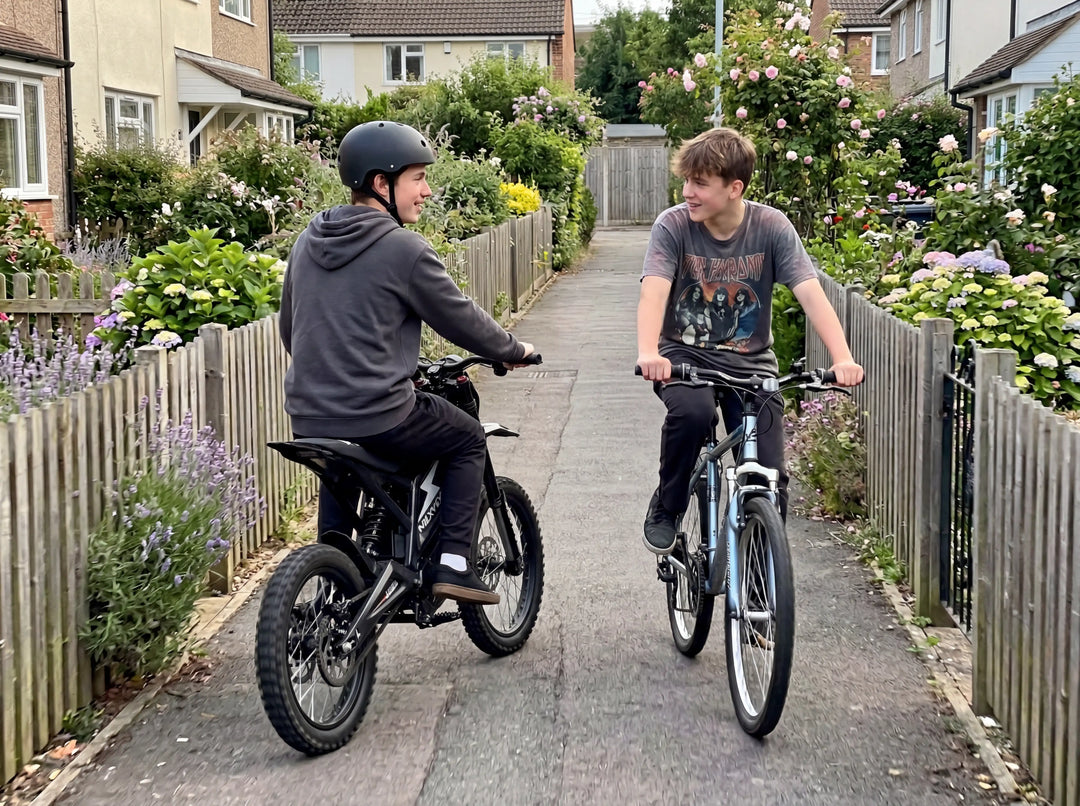 A teenager on a black NEXVEY FLASH electric dirt bike riding alongside a friend on a bicycle through a narrow flowering residential alleyway