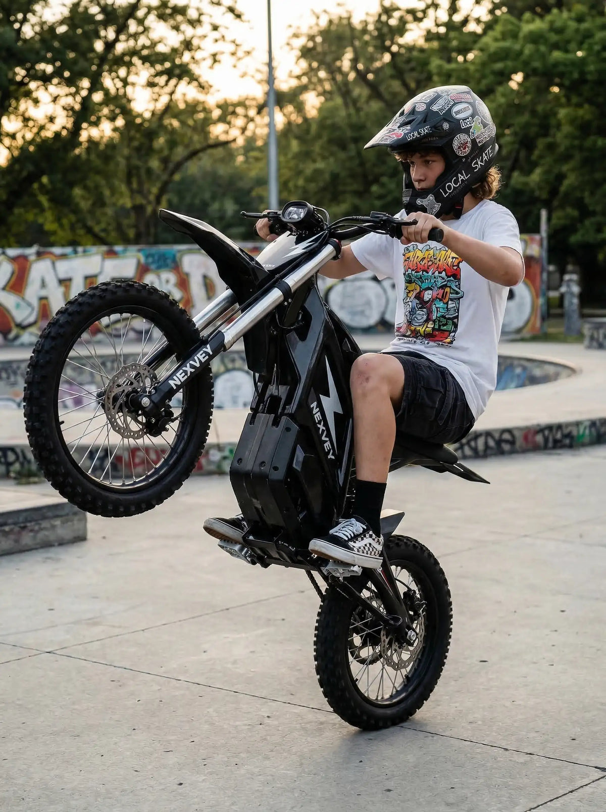 A young rider wearing a full-face helmet performing a wheelie on a NEXVEY electric dirt bike at a concrete skatepark during the day
