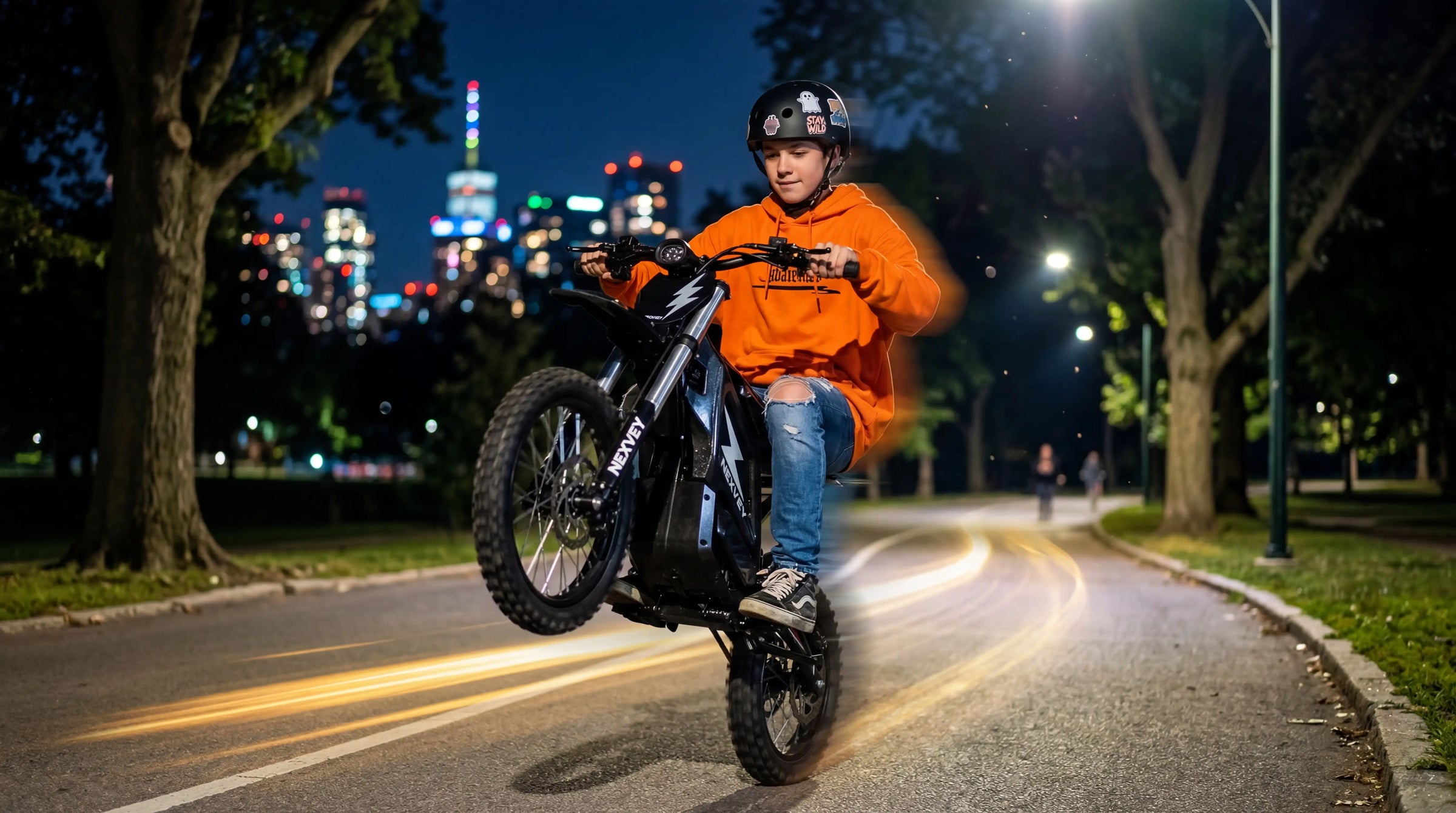 A person in an orange hoodie and black helmet performing a wheelie on an electric dirt bike at night in a park, with city lights and light streaks in the background