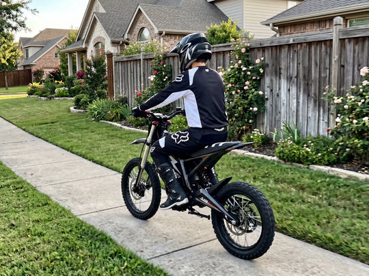 Rear view of a rider in professional black and white racing gear on a black NEXVEY FLASH electric dirt bike, cruising along a clean suburban sidewalk lined with green lawns and flowers