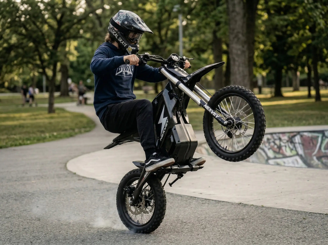 A rider in a blue hoodie and sticker-covered helmet performing a powerful wheelie on a black NEXVEY FLASH electric dirt bike with tire smoke at a graffiti skatepark