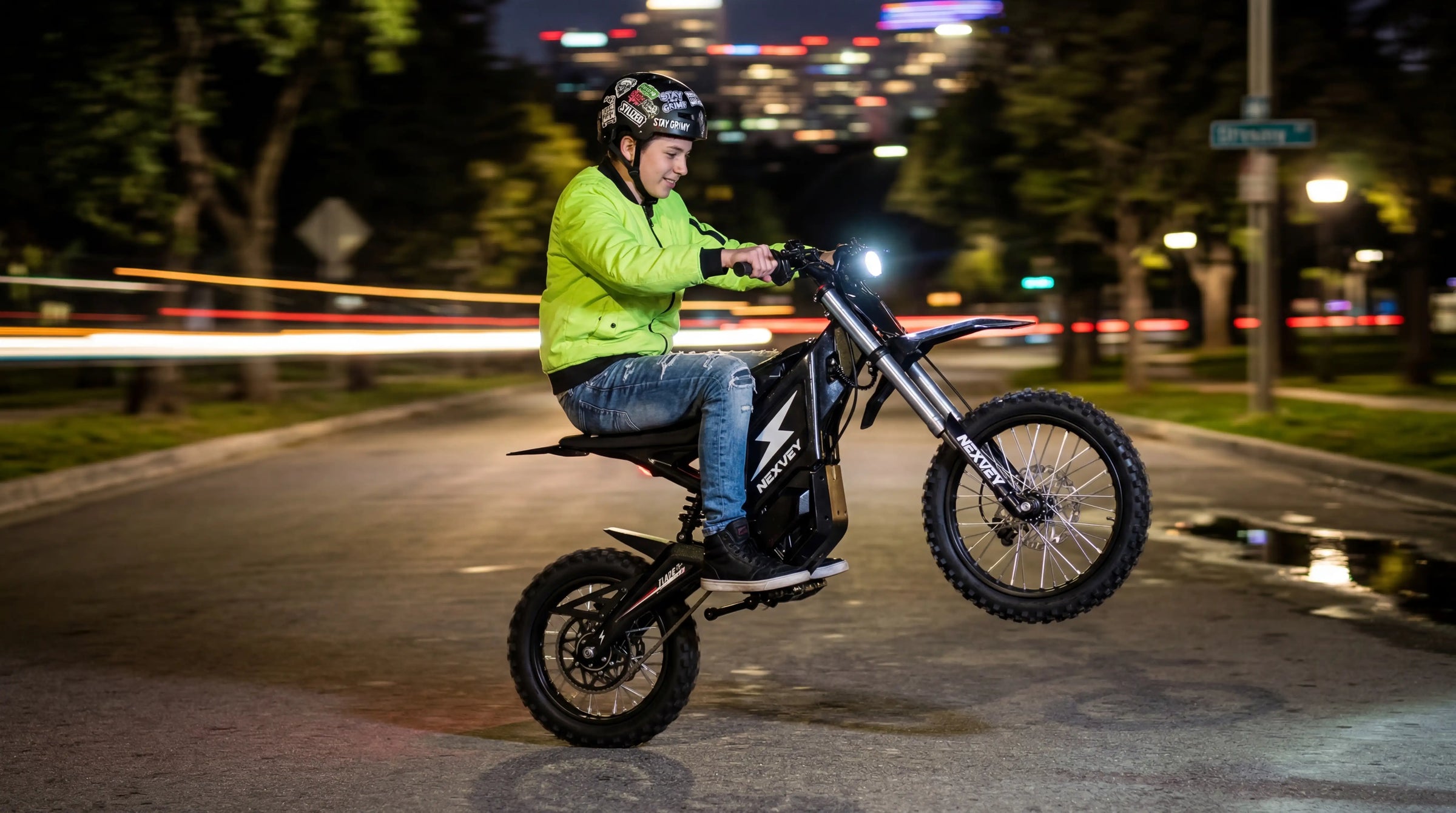 A young rider in a lime green jacket performing a wheelie on a NEXVEY electric dirt bike at night with blurred city lights.