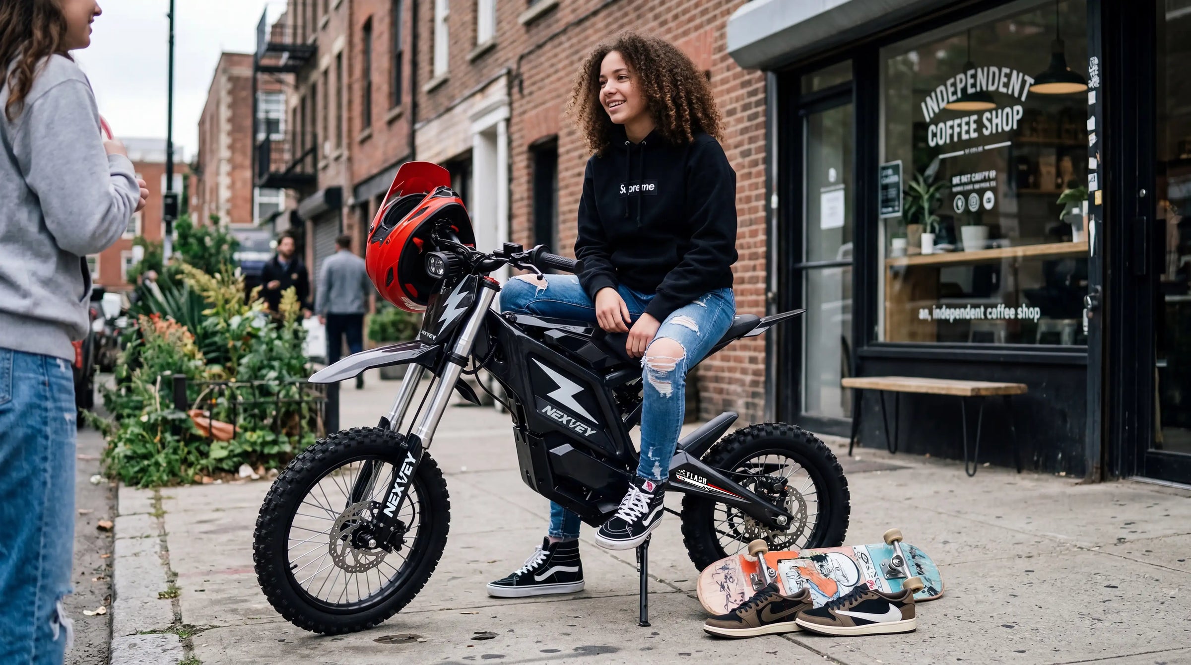 A stylish teenage girl with curly hair sitting on a Nexvey black electric dirt bike on a city sidewalk, featuring a red helmet and skateboards for a modern youth lifestyle