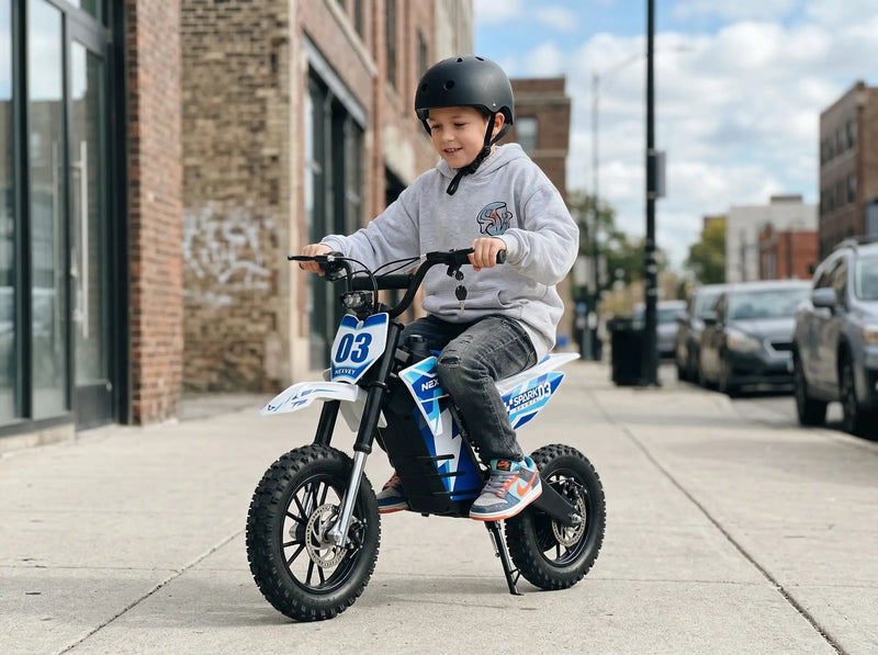 A young boy wearing a black safety helmet riding a NEXVEY Spark D3 blue and white electric dirt bike on a city sidewalk