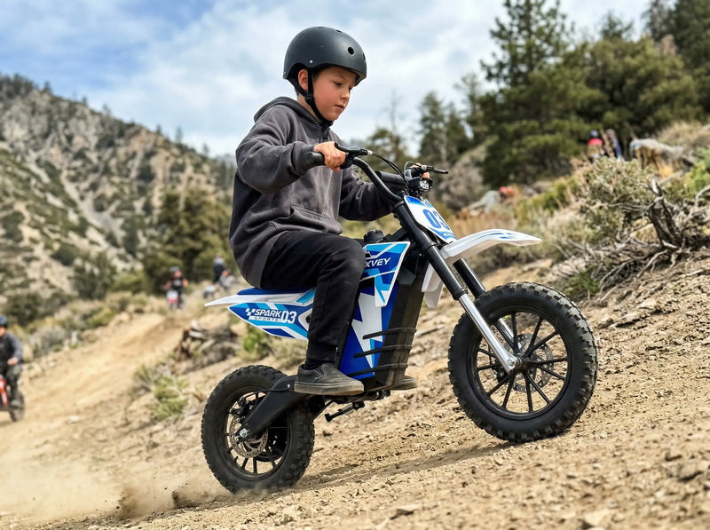 A young boy riding a NEXVEY Spark D3 electric dirt bike uphill on a rugged mountain trail with pine trees in the background