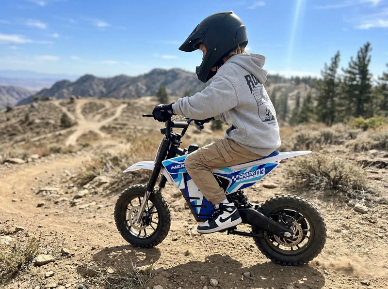 Young boy wearing a safety helmet riding a NEXVEY Spark D3 blue electric dirt bike on an off-road mountain trail.