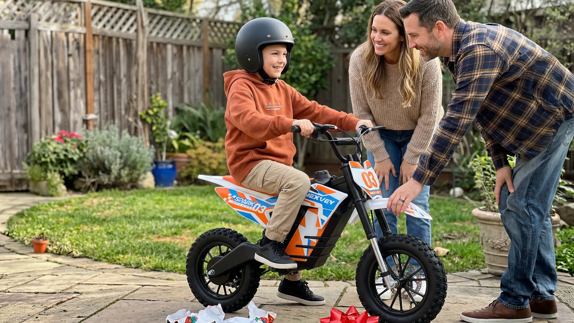 A happy young boy wearing a helmet sits on a NEXVEY electric dirt bike while his parents smile in a backyard setting